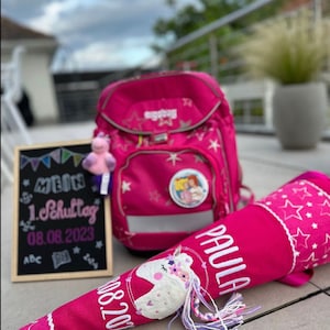 Könnte beinhalten: Ein pinkfarbener Schulrucksack, eine passende kegelförmige Zuckertüte und eine kleine Tafel. Der Kegel ist mit Sternen und einem Einhorn verziert. Auf der Tafel steht "Mein 1. Schultag 08.08.2023". Ein kleines Stofftier hängt am Rucksack.