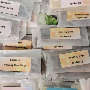 May include: An assortment of wrapped soap samples, each labeled with a unique scent name. The labels include "Vanilla Latte," "Moonlit Skies," and "Cowboy Beer Soap." The samples are packaged in small, clear wrappers.