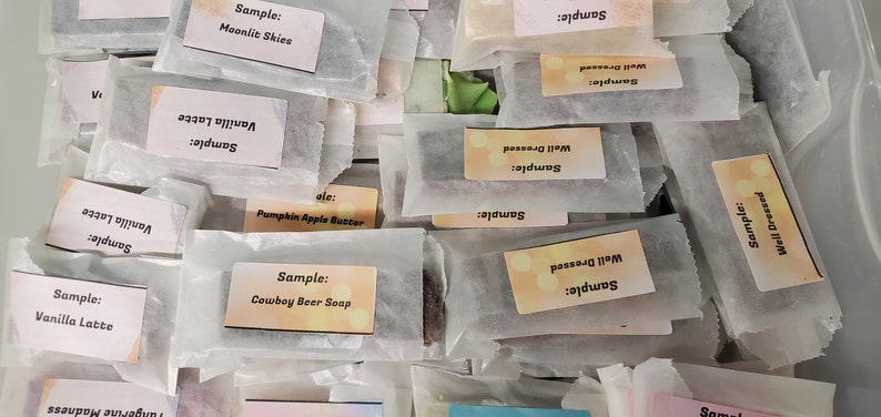 May include: An assortment of wrapped soap samples, each labeled with a unique scent name. The labels include "Vanilla Latte," "Moonlit Skies," and "Cowboy Beer Soap." The samples are packaged in small, clear wrappers.