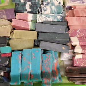 May include: A collection of handmade soap bars in various colors and designs. The soaps are rectangular and feature colors like purple, pink, green, blue, and black. Some bars have decorative patterns and embedded elements.