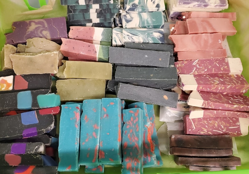 May include: A collection of handmade soap bars in various colors and designs. The soaps are rectangular and feature colors like purple, pink, green, blue, and black. Some bars have decorative patterns and embedded elements.