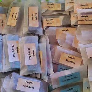 May include: A collection of wrapped soap samples in a green basket. Each sample is labeled with a scent name, such as "Cheeky Teal," "Bay Rum," and "Rose Clay." The samples are in small, rectangular shapes and wrapped in white paper.