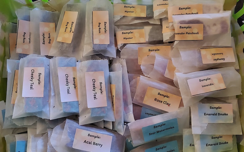 May include: A collection of wrapped soap samples in a green basket. Each sample is labeled with a scent name, such as "Cheeky Teal," "Bay Rum," and "Rose Clay." The samples are in small, rectangular shapes and wrapped in white paper.