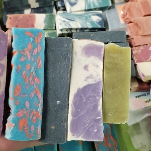 May include: A variety of handmade soap bars in various colors and designs. The bars include shades of purple, blue, green, and pink, with decorative elements like gold accents and floral patterns. The soaps are rectangular and appear to be freshly cut.
