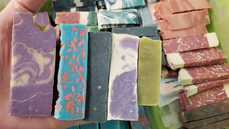May include: A variety of handmade soap bars in various colors and designs. The bars include shades of purple, blue, green, and pink, with decorative elements like gold accents and floral patterns. The soaps are rectangular and appear to be freshly cut.