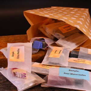 May include: A brown paper bag with white polka dots, overflowing with small, wrapped soap samples. The samples are labeled with various scents, including "Citrus Medley" and "Sour Watermelon". The soaps are in various colors.