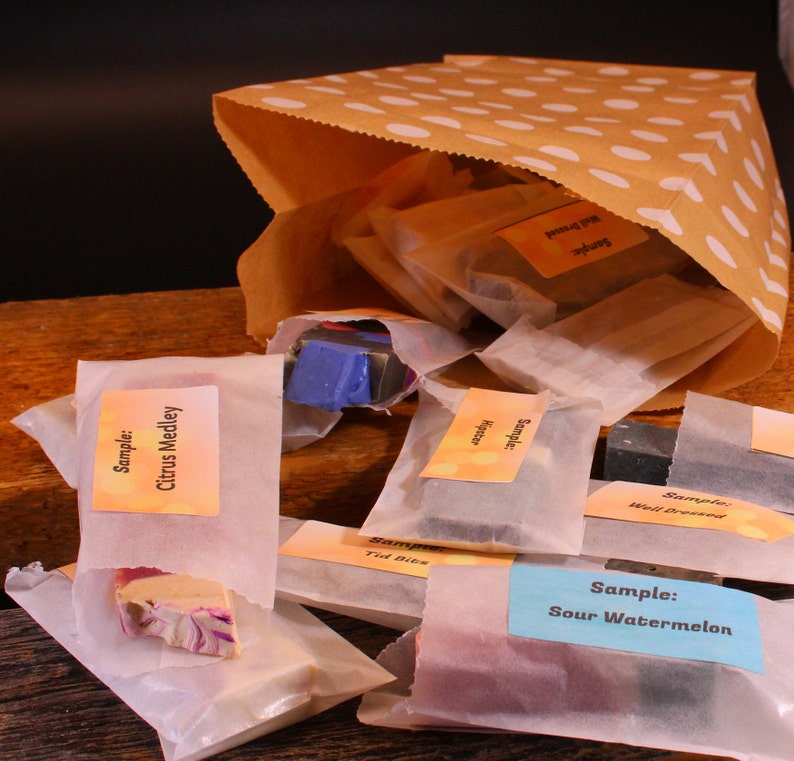 May include: A brown paper bag with white polka dots, overflowing with small, wrapped soap samples. The samples are labeled with various scents, including "Citrus Medley" and "Sour Watermelon". The soaps are in various colors.