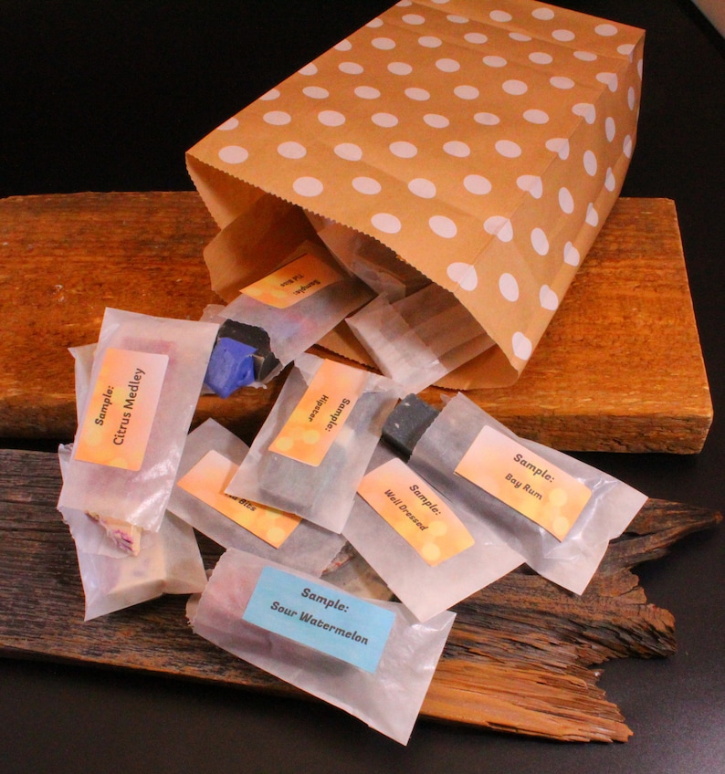 May include: A brown paper bag with white polka dots is tipped over, spilling out small, wrapped samples. The samples are labeled with various scents, including "Citrus Medley," "Sour Watermelon," and "Bay Rum." The bag rests on a wooden surface.