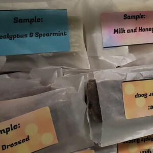 May include: Assortment of wrapped soap samples with colorful labels. Labels include "Eucalyptus & Spearmint", "Milk and Honey", "Tangerine Madness", "Vanilla Latte", "Cowboy Beer Soap", and "Well Dressed". The soaps are packaged in white paper.