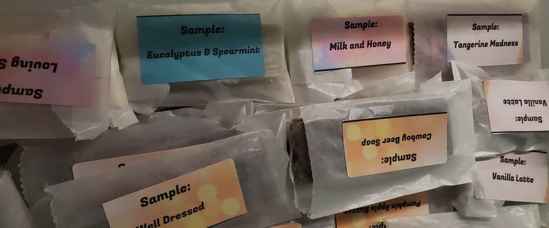 May include: Assortment of wrapped soap samples with colorful labels. Labels include "Eucalyptus & Spearmint", "Milk and Honey", "Tangerine Madness", "Vanilla Latte", "Cowboy Beer Soap", and "Well Dressed". The soaps are packaged in white paper.