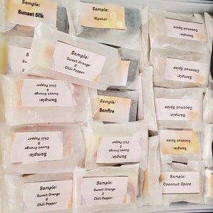 May include: A collection of wrapped soap samples, each labeled with a unique scent name. The samples are in a variety of colors, including orange, green, and white. The labels include scents like "Sweet Orange & Chili Pepper" and "Coconut Spice."