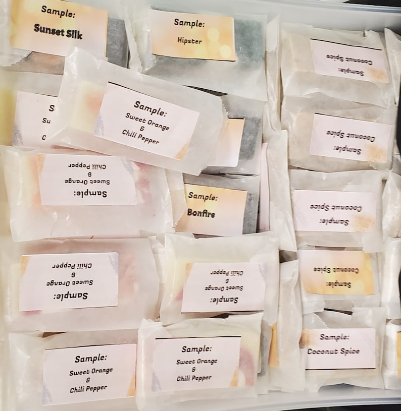 May include: A collection of wrapped soap samples, each labeled with a unique scent name. The samples are in a variety of colors, including orange, green, and white. The labels include scents like "Sweet Orange & Chili Pepper" and "Coconut Spice."