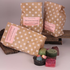 Peut inclure: Trois sacs en papier brun à pois blancs, chacun portant une étiquette rose indiquant Desert Lily Soaps Odds & Ends Soap Scraps. Plusieurs petits morceaux de savon multicolores de formes et textures variées sont disposés devant les sacs.