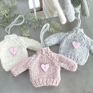 May include: Four knitted sweater ornaments with heart-shaped buttons. The sweaters are in white, pink, grey, and green.
