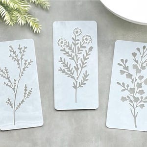 May include: Three white plastic stencils with floral designs. The stencils are rectangular and feature different floral patterns, including a single stem with leaves and a cluster of flowers.