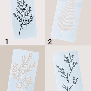 May include: Four rectangular stencils featuring botanical designs. The stencils are white with plant illustrations in grey and beige. The size is indicated as 15x7.5cm. The designs include fern and branch patterns.