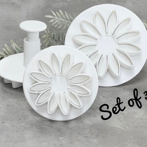 May include: Set of three white plastic daisy cookie cutters. The cutters are different sizes and have a plunger for easy use. The text "Set of 3" is written on the surface.