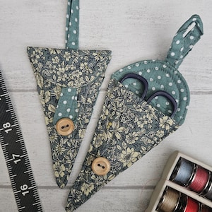 May include: Two triangular fabric scissor sheaths featuring a floral pattern in shades of blue, green, and cream, accented with a teal and white polka dot fabric, a wooden button closure, and a hanging loop, shown with a ruler and thread spools.