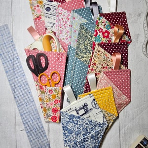 May include: A selection of patterned fabric scissor holders, featuring polka dots and floral prints. Each holder has a ribbon loop and holds sewing tools such as scissors and tape. A ruler and measuring tape are also present.