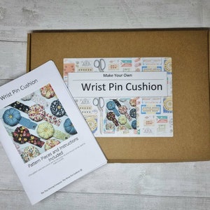 May include: A craft kit for making wrist pin cushions. The kit includes a booklet titled "Wrist Pin Cushion" with pattern pieces and instructions, and a cardboard box with a label that reads "Make Your Own Wrist Pin Cushion."