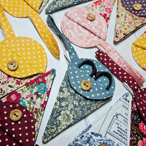 May include: A collection of fabric scissor cases in diverse patterns and colours. Designs include polka dots, floral prints, and solid hues. Each case features a wooden button fastening and a hanging loop.