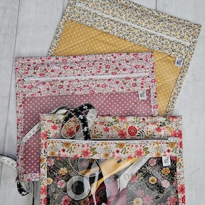 May include: Three rectangular quilted zip pouches with floral patterns in yellow, pink, and black. Each pouch features a clear vinyl front panel, a white zip, and a small tag labelled Tiny. A black and white measuring tape is coiled on top.