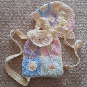 May include: A crocheted backpack and hat set in pastel colours with daisy flower designs. The backpack has a white strap and a button closure. The hat has a white brim and a crocheted flower design.