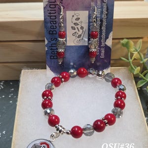 May include: A red and gray beaded bracelet and matching earrings set. The bracelet features a charm with the Ohio State Buckeyes logo. The earrings and bracelet are displayed on a small box with a tag that reads "Beth's Beadtique".