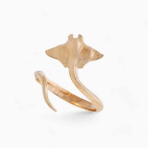 May include: A gold-coloured ring with a manta ray design. The ring wraps around the finger, with the ray's body forming the top and its wings extending outwards. The ring is made of a shiny, metallic material.