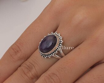 Op de afbeelding: Een zilveren ring met een grote, ovaalvormige, dieppaarse amethist edelsteen in een decoratieve zetting. De zetting is omringd door een kralen, geschulpt randontwerp. De ringband splitst zich aan de basis in twee dunne, gepolijste zilveren schachten.