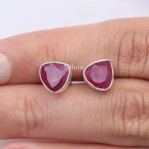 Puede incluir: Un par de pendientes de botón con piedras preciosas de rubí rojo de forma triangular y facetadas, engastadas en metal de tono plateado. Las piedras tienen un tono carmesí profundo con inclusiones internas visibles y un engaste metálico.
