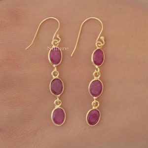 May include: Gold-tone drop earrings featuring three oval-cut, deep red gemstones. The earrings have a simple, elegant design with a hook closure. The gemstones are set in a gold bezel setting, creating a classic and timeless look.