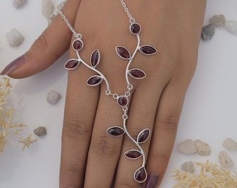 Puede incluir: Una cadena de mano plateada con una delicada estructura en forma de enredadera adornada con múltiples piedras preciosas de granate rojo oscuro talladas en marquise. La cadena de metal se ramifica en monturas con forma de hoja.
