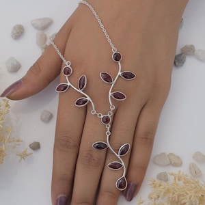 May include: A silver necklace with a delicate branch design featuring red gemstones. The necklace is connected to a ring that can be worn on the finger.
