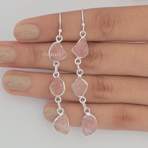 May include: A pair of drop earrings featuring three raw, light pink stones set in silver-toned metal frames. Each earring consists of three stones connected by silver jump rings, suspended from a silver hook. The text Sillure is visible.