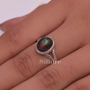 May include: A silver ring featuring an oval-shaped dark stone with iridescent flashes of red and green. The band has a split design, and the word Sillure is printed in white text below the ring setting on the image surface.