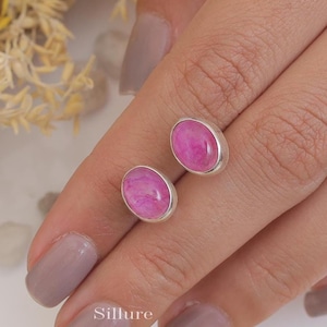 Peut inclure: Une paire de boucles d'oreilles en argent avec des pierres ovales roses. Les boucles d'oreilles sont présentées sur la main d'une personne.