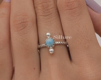 May include: A silver ring featuring a central round light blue stone with a marbled texture, flanked by two small silver spheres. The band has a twisted rope design. The word Sillure is printed in white text to the right of the stone.