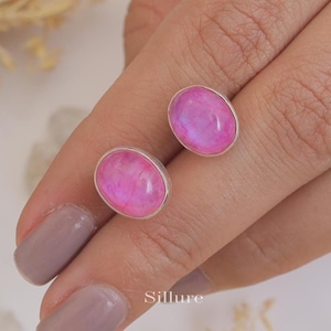 Puede incluir: Un par de pendientes de plata con piedras ovaladas de color rosa. Las piedras tienen un acabado brillante e iridiscente.