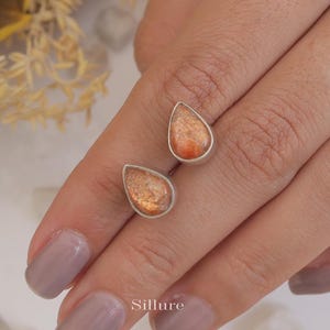 May include: A pair of silver teardrop earrings with orange sunstone gemstones. The earrings are shown on a hand.