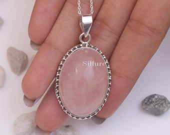 May include: A silver pendant necklace featuring a large, smooth, oval-shaped rose quartz gemstone set in a beaded silver frame. The pendant hangs from a delicate silver chain. The word Sillure is printed in white text on the surface of the stone.