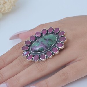 Op de afbeelding: Een zilveren ring met een grote ovale groene en paarse edelsteen omringd door een halo van kleinere roze edelstenen.