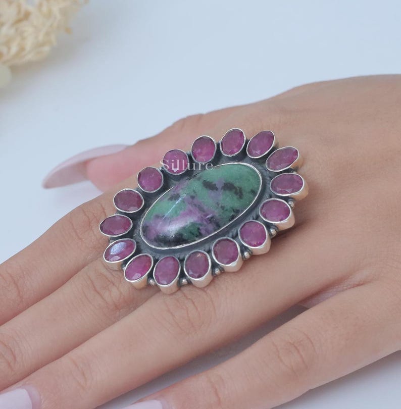 Op de afbeelding: Een zilveren ring met een grote ovale groene en paarse edelsteen omringd door een halo van kleinere roze edelstenen.