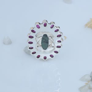 Op de afbeelding: Een zilveren ring met een groene ovale edelsteen omringd door een cirkel van kleine roze edelstenen. De ring is gegraveerd met het woord "Silure".