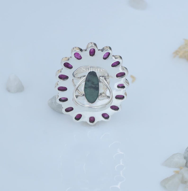 Op de afbeelding: Een zilveren ring met een groene ovale edelsteen omringd door een cirkel van kleine roze edelstenen. De ring is gegraveerd met het woord "Silure".