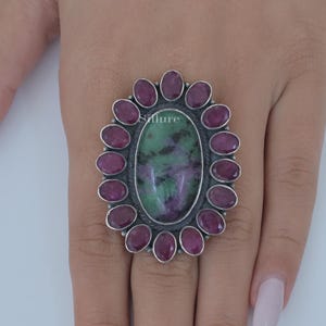 Op de afbeelding: Een zilveren ring met een grote ovale groene en paarse edelsteen omringd door een halo van kleinere roze edelstenen.