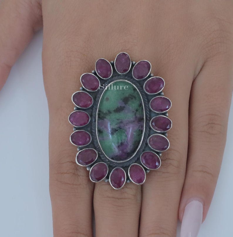 Op de afbeelding: Een zilveren ring met een grote ovale groene en paarse edelsteen omringd door een halo van kleinere roze edelstenen.