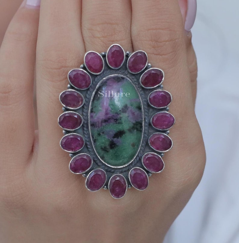 Op de afbeelding: Een zilveren ring met een grote ovale groene en paarse edelsteen, omringd door een halo van kleinere roze edelstenen. De ring wordt aan een vinger gedragen.