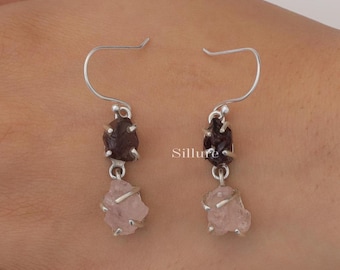 May include: A pair of drop earrings featuring silver-toned hooks, each suspending a dark red raw gemstone above a pale pink raw gemstone. The stones are held in place by thin silver-toned wire settings. The word Sillure is printed in the center.