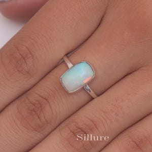 May include: A silver ring featuring a rectangular opal gemstone. The opal is a milky white colour with flashes of blue and green.